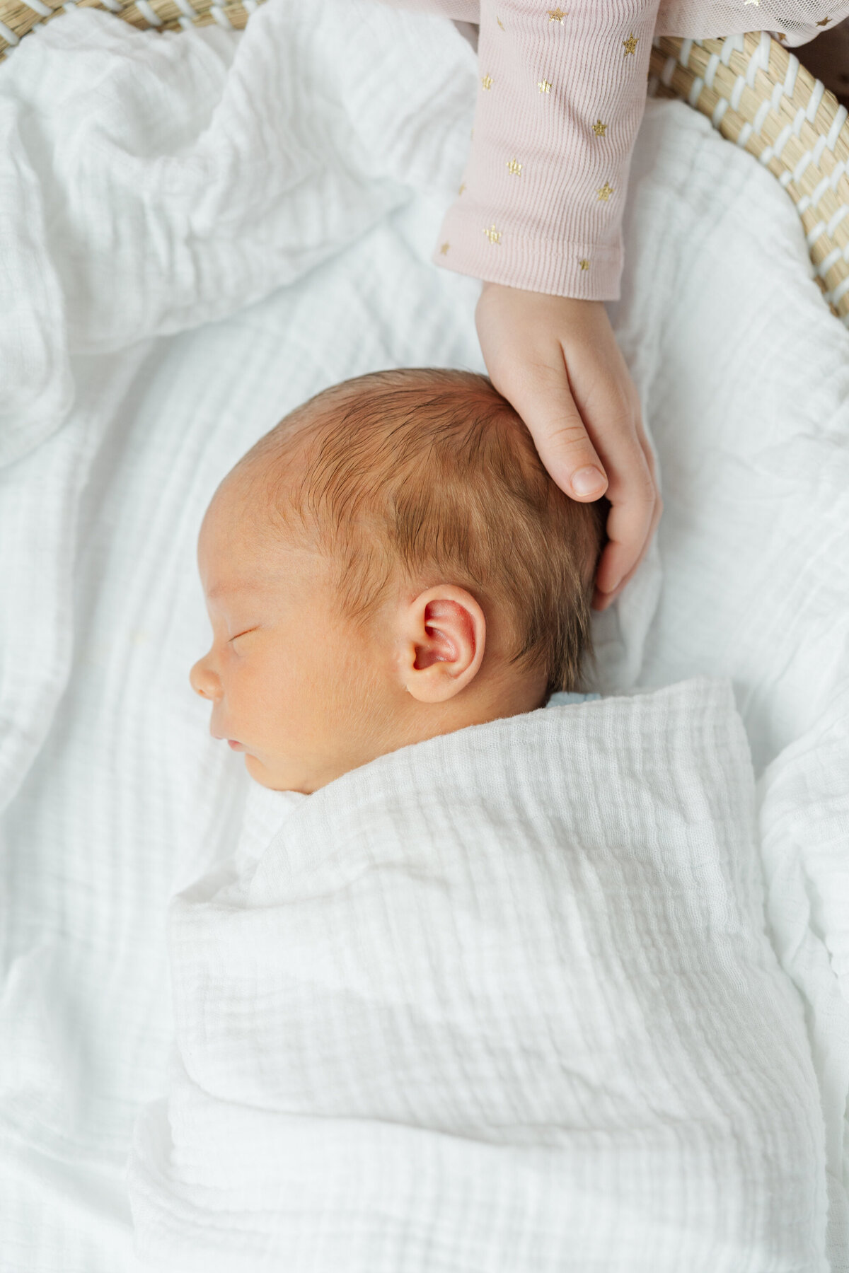 Newborn baby peacefully lying on their side with an older sibling's hand gently resting on their head, showing a sweet bond