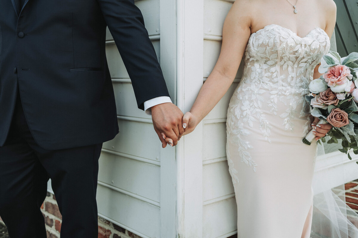 Close-up of a bride and groom sharing a first touch around a corner at the Burlington Weddings and Event Venue in Virginia. The bride, wearing a lace wedding dress and holding a bouquet of roses, and the groom in a navy suit hold hands. Shot by Quinn Photography LLC.