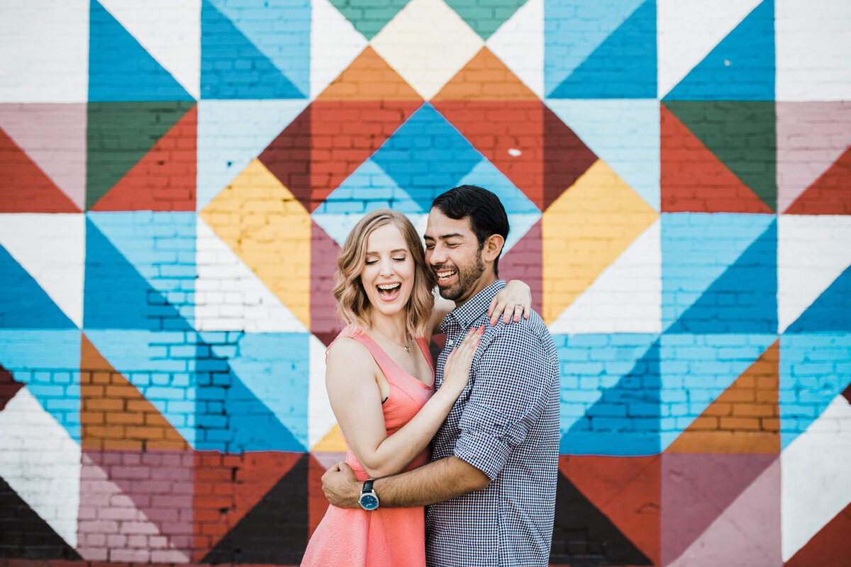 A couple holding each other close and laughing in front of a colorful, geometric mural during their engagement session in Deep Ellum in Dallas, Texas. The woman on the left is wearing a sleeveless salmon dress while the man on the right is wearing a striped dress shirt.