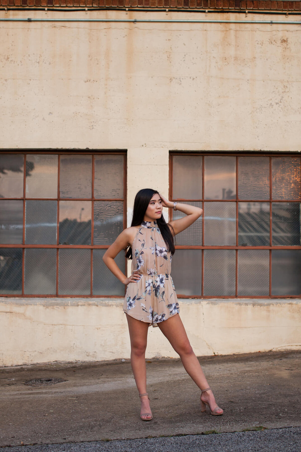 A confident Asian teen poses for her senior pictures in front of some industrial looking windows in a downtown city parking lot. Captured by Springfield, MO senior photographer Dynae Levingston.
