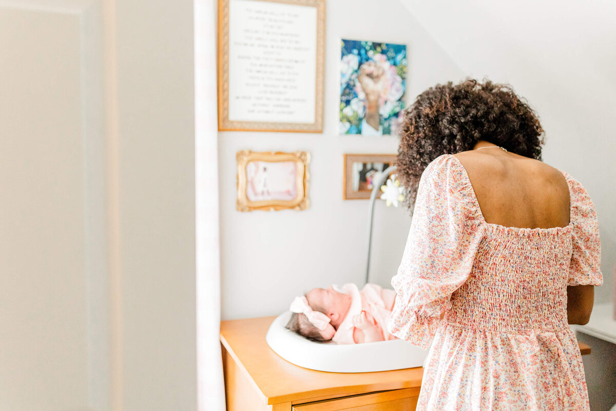 Seen through a doorway, a mother in a pink dress changes her newborn daughter's diaper