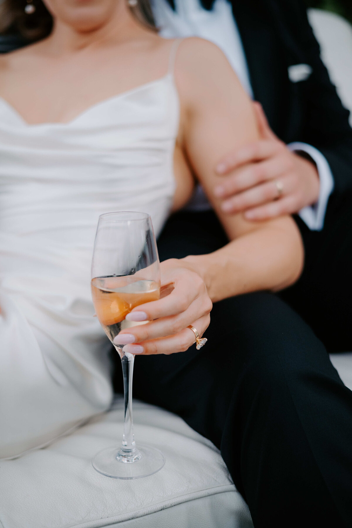 Close-up of bride in a satin gown holding a champagne glass, with the groom’s arm around her, showcasing wedding rings