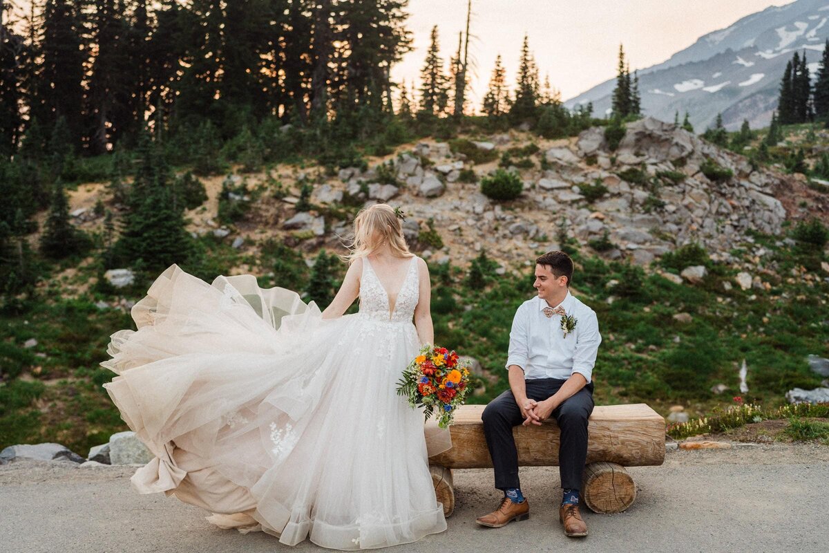 a bride throwing her dirty dress as her groom watches her from a bench during their Mount Rainier Elopement captured by Washington Elopement Photographer Sage Trails.