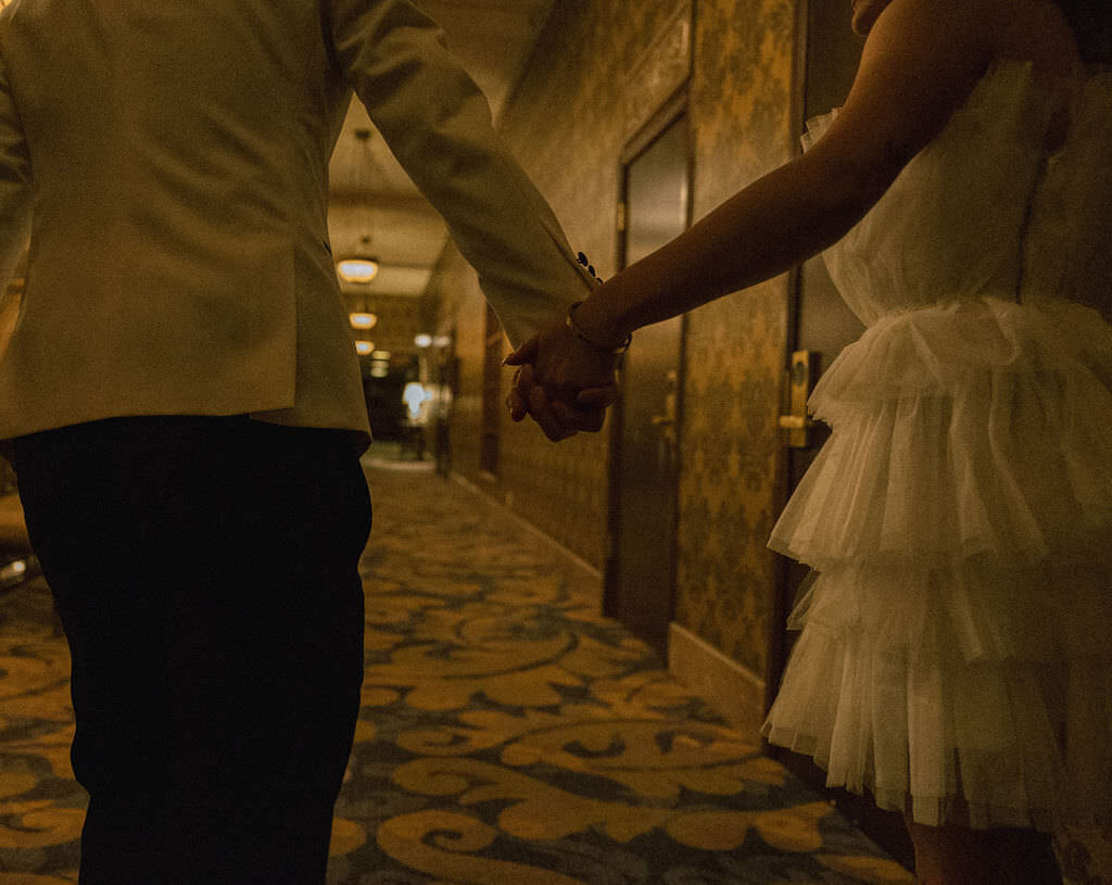 A wedding couple holding hands and walking down a hallway.