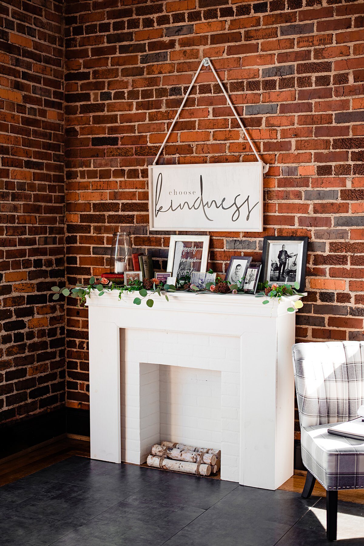 Set against an exposed brick wall is a white mantle with birchwood logs. Above the mantle is a sign that says KINDNESS. Set on the mantle are antique books, a large glass hurricane lantern with a white pillar candle and an assortment of old photographs for a wedding memorial table. Off to the right is a white and gray plaid sofa.