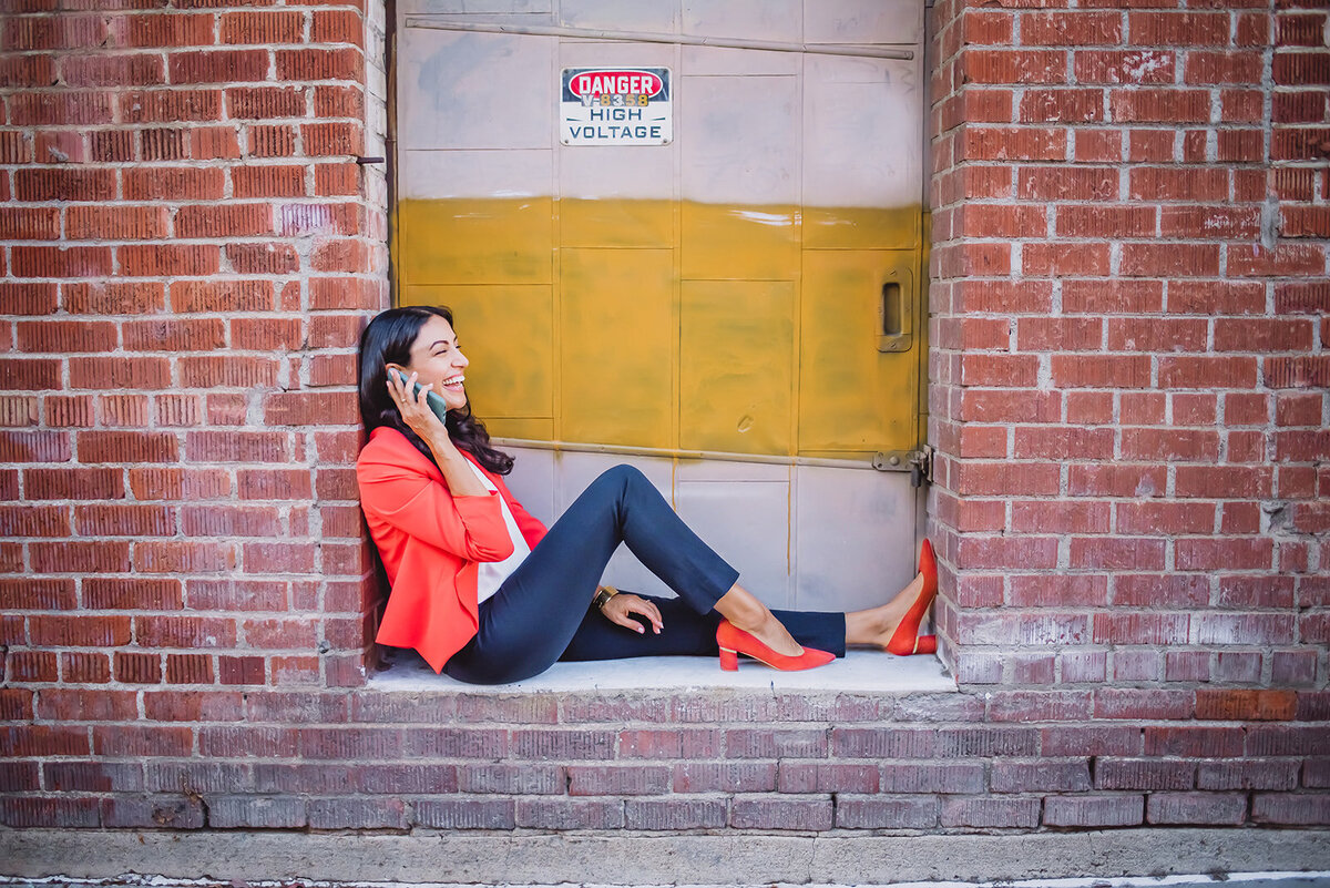 A woman in an orange blazer sits and laughs during a phonecall.
