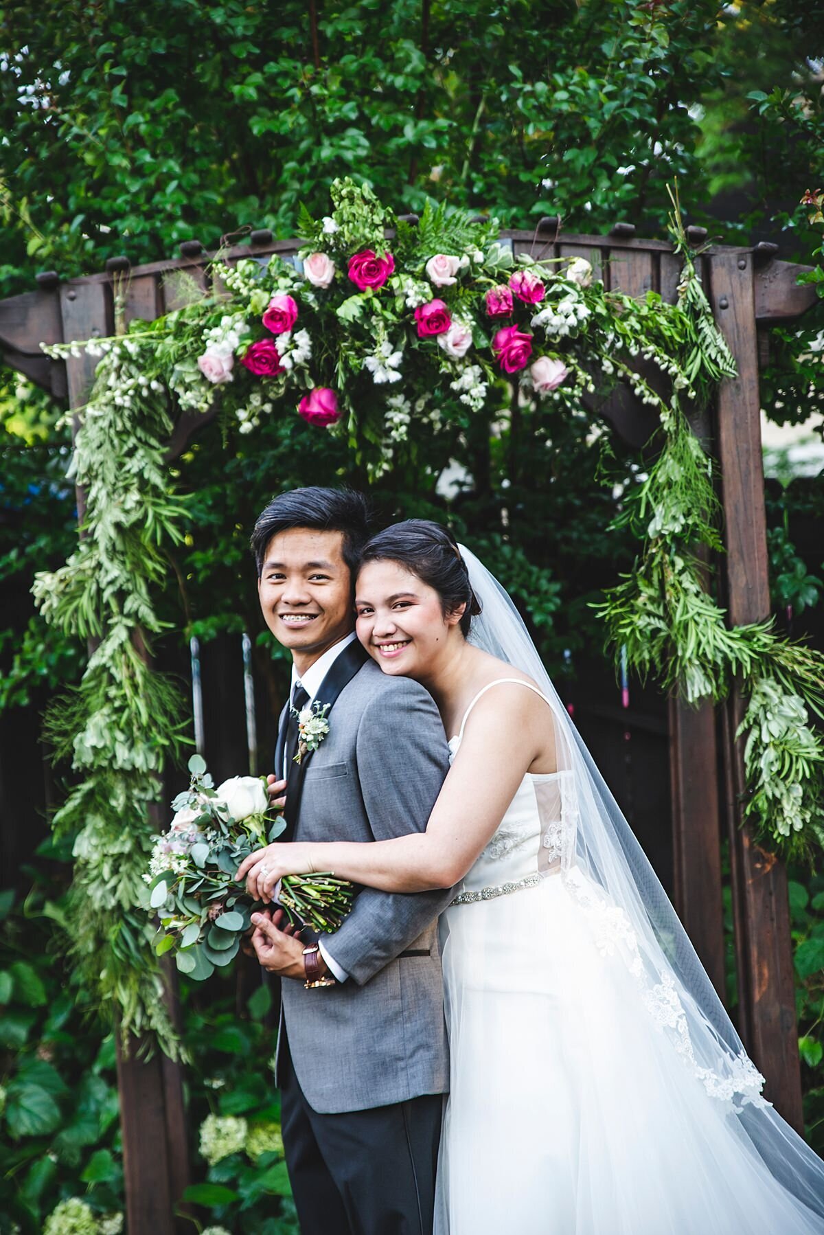 The bride, wearing a flowing wedding dress and long veil embraces the groom who is wearing a tuxedo with a grey jacket and white boutonniere. The bride is holding a large bouquet of white roses, white peonies, white carnations, white tea roses and silver dollar eucalyptus. They are standing in front of a wooden arbor decorated with a garland of evergreen, white roses and pink roses.
