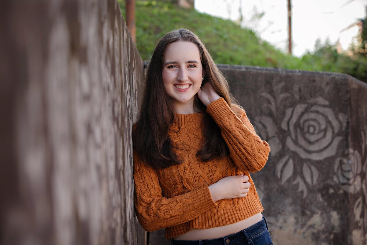 Beautiful HS senior in orange sweater leaning against concrete wall with faded stamped rose designs at Camp North End Charlotte NC