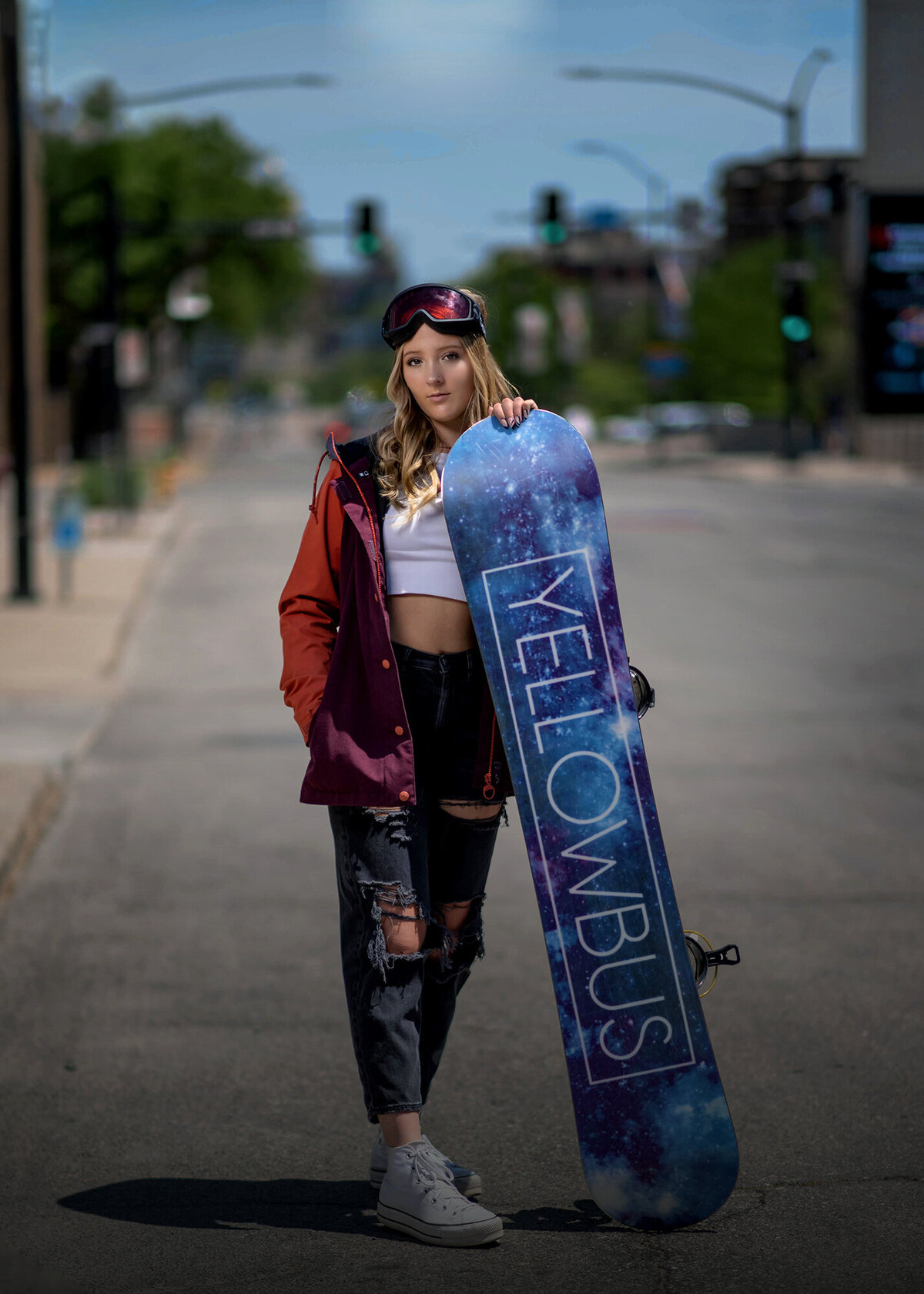A girl leans on her blue snowboard in pants and red hoodie in the street