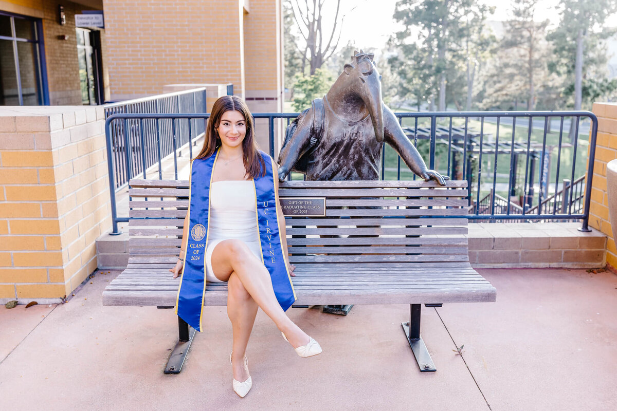 A woman in a white dress and blue graduation stole sits on a bench next to a bronze anteater statue. She is smiling, with one arm resting on her knee. Trees and a building are visible in the background.