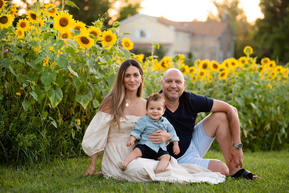 mom and dad sitting on the ground with their baby boy in front of a sunflower field in the golden hour for their summer Burlington family photographer session