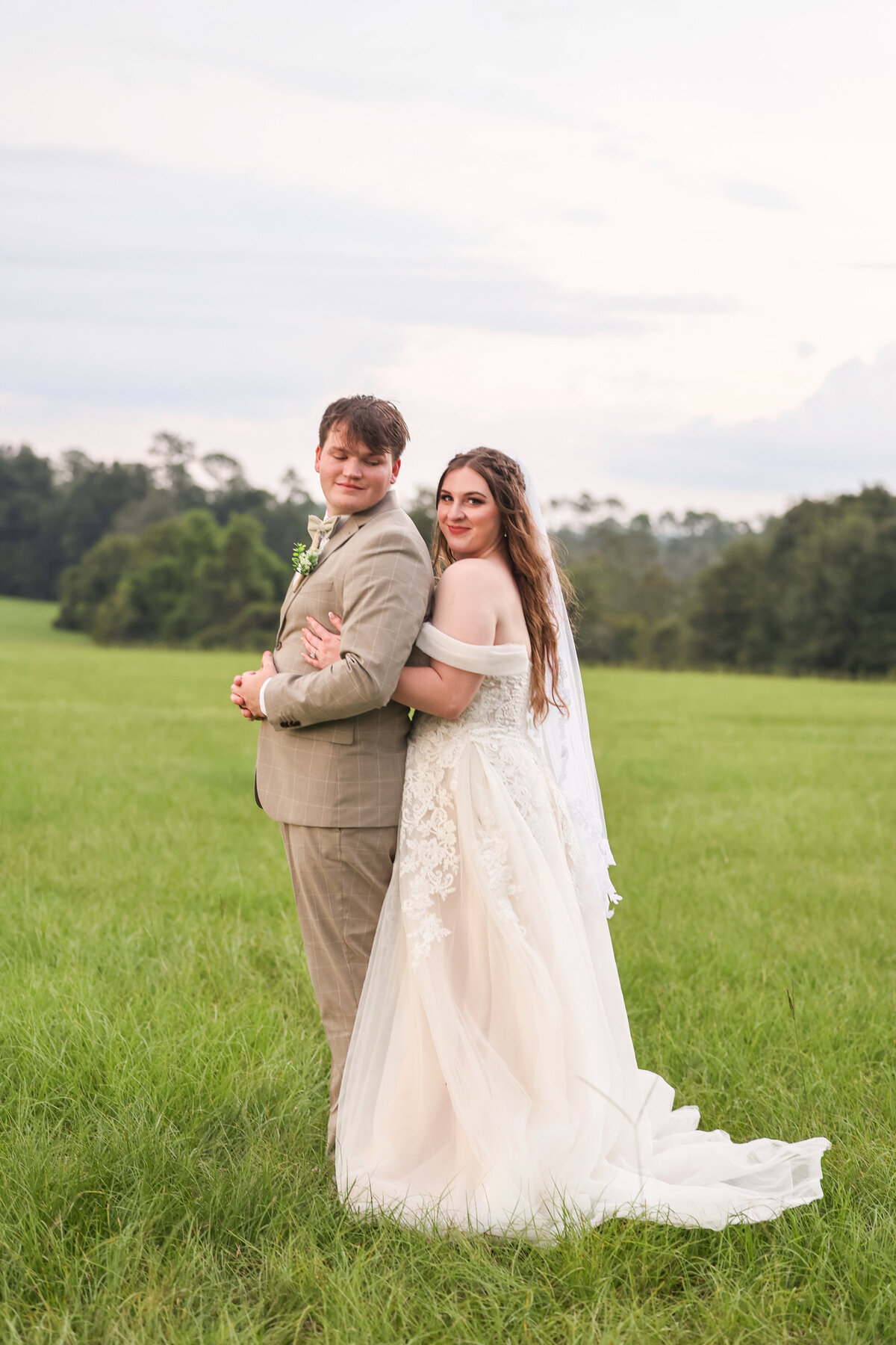 groom wearing light brown suit looking over shoulder at his bride wearing white tulle dress in grassy area