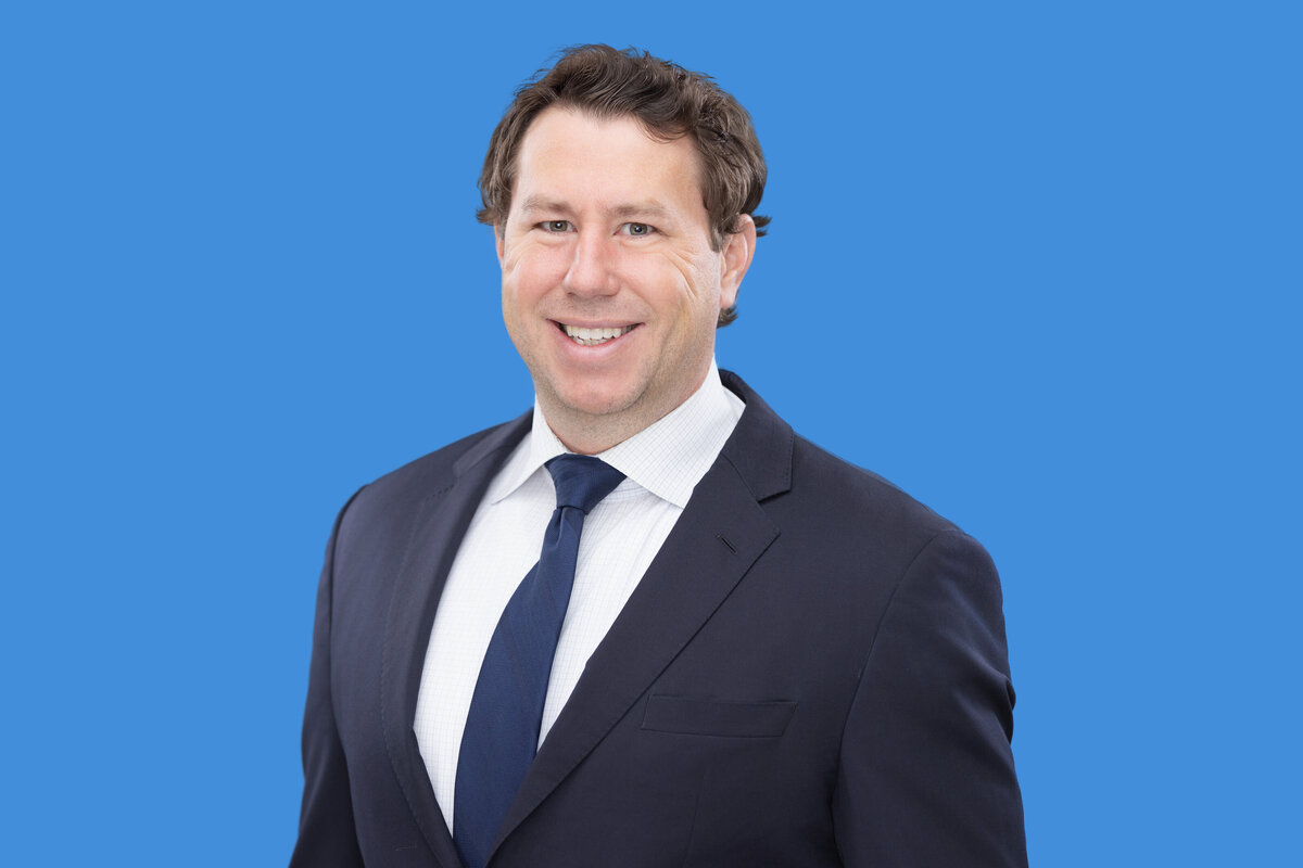 Professional headshot of a confident gentleman against a bright blue background in Cincinnati. He sports a sharp black suit, blue tie, and a white shirt. His warm smile and approachable demeanor stand out, making this an ideal representation for corporate profiles.