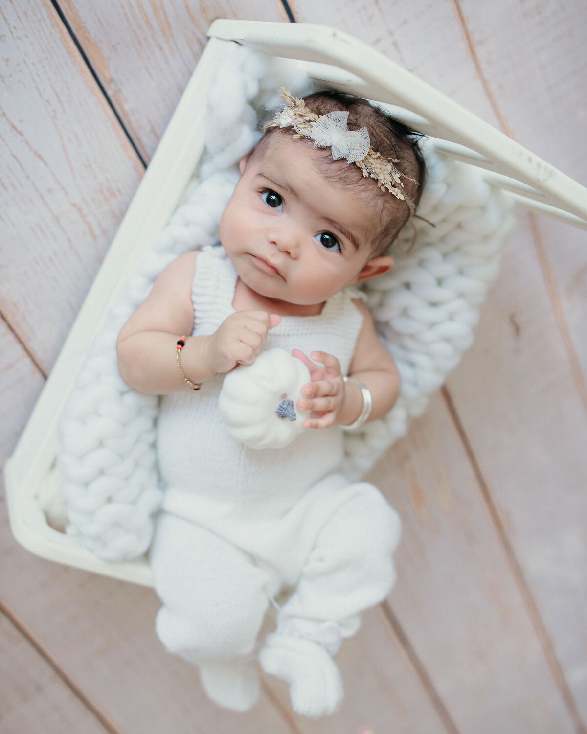 Newborn girl awake and looking right at camera dressed in white long pants romper holding white mini pumpkin and laying in white baby bed