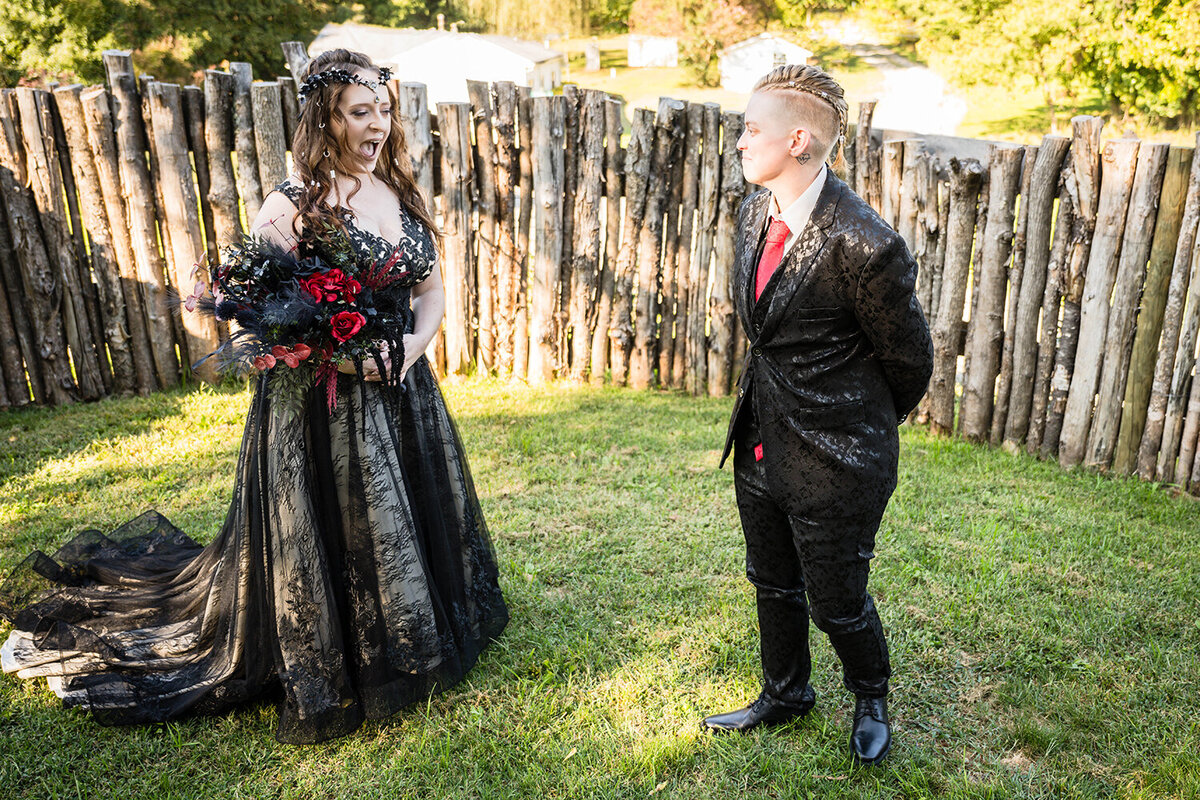 Two queer marriers stand in the backyard to be set up for their first look at an Airbnb they rented in Roanoke, Virginia on their elopement. One marrier, wearing a black floral suit, turns towards her significant other, who is wearing a dress and holding a bouquet. The marrier wearing a black dress has their mouth open and their expression is surprised.