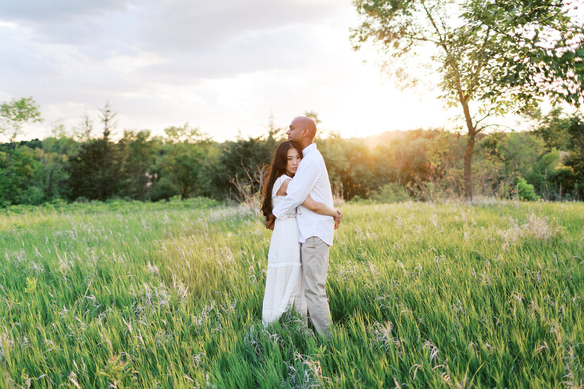Two people in all-white attire stand in the tall grass of Elm Creek Park in Maple Grove at sunset, their hair glowing as they share a silent, romantic embrace with sun rays showering them, captured by a Minnesota wedding photographer.