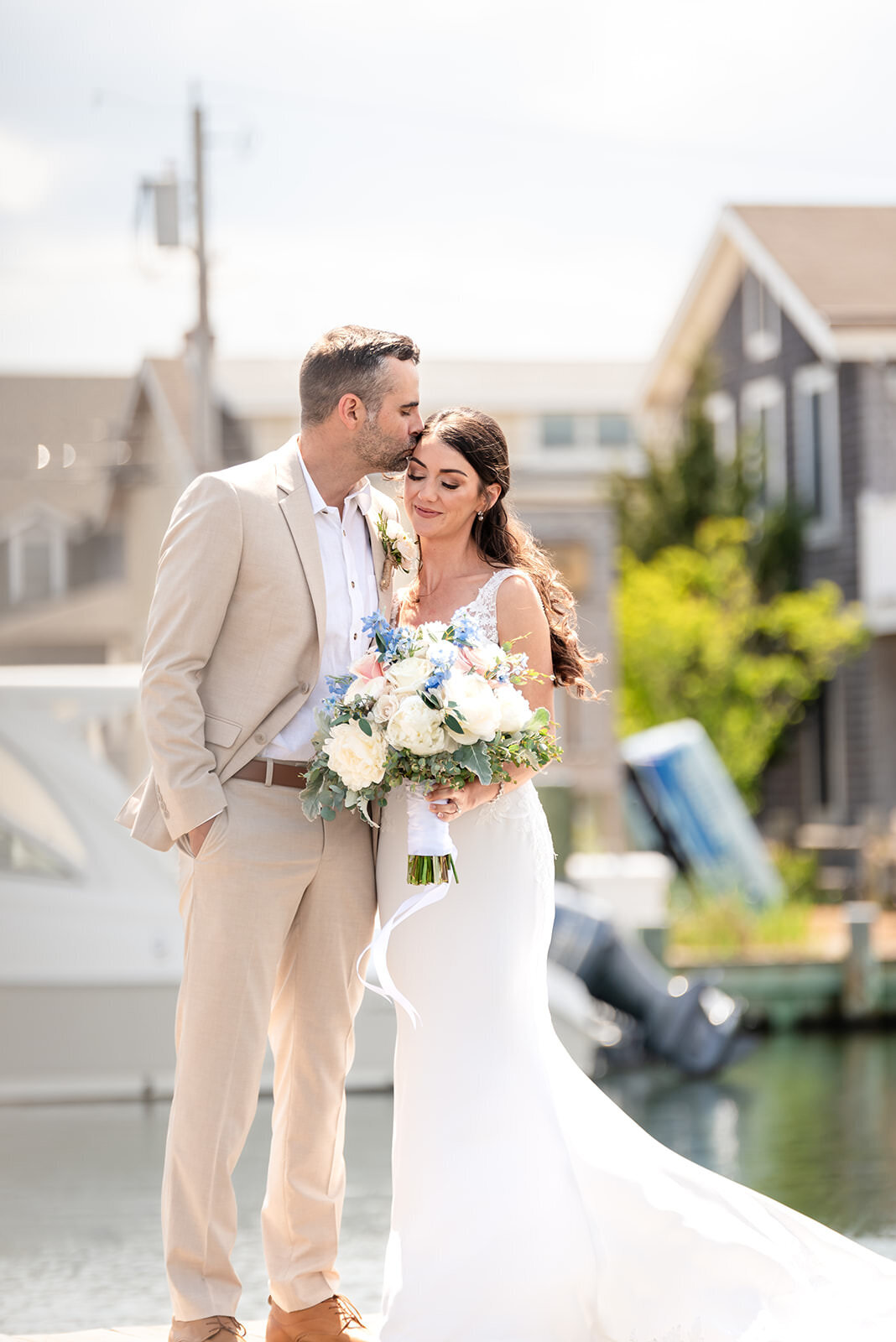 The couple stands close together on a dock, with the groom kissing the bride's forehead while she holds a bouquet of white and blue flowers.