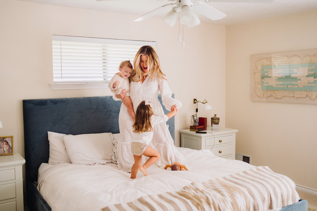 A mom with her daughters on a white and blue bed. The woman, dressed in a long white dress, is holding the baby while gently lifting the older girl who is looking up. The background features an aquamarine painting, a bedside table with a lamp, and perfumes