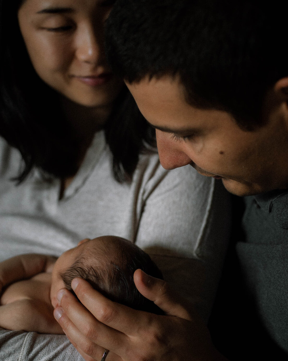 Father gently holding newborn’s head while mother looks on during intimate family session.