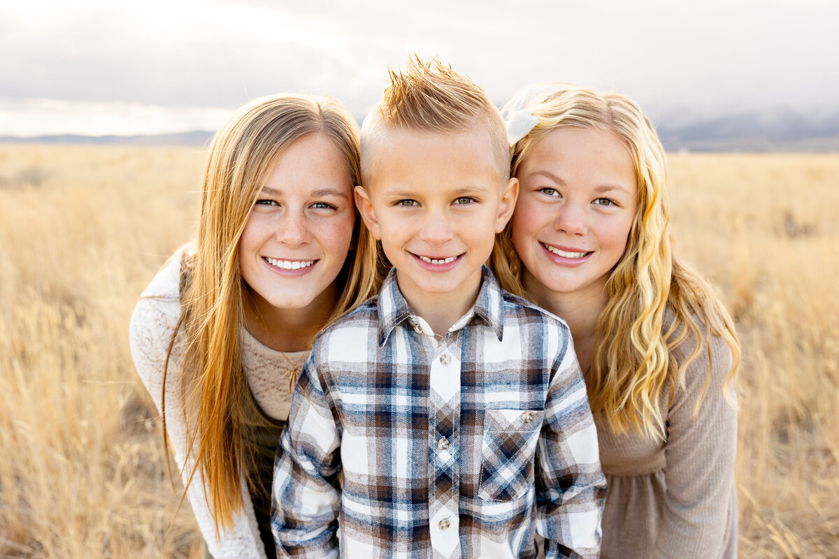 Three siblings—two sisters and a younger brother—cuddling close together and smiling at the camera. The golden grass and distant mountains create a warm and dreamy backdrop.