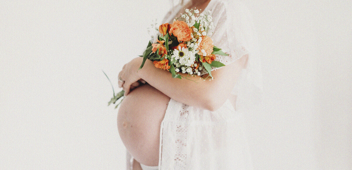 Minimalist studio shot of a woman in a sheer robe holding a bright floral bouquet against her bump.