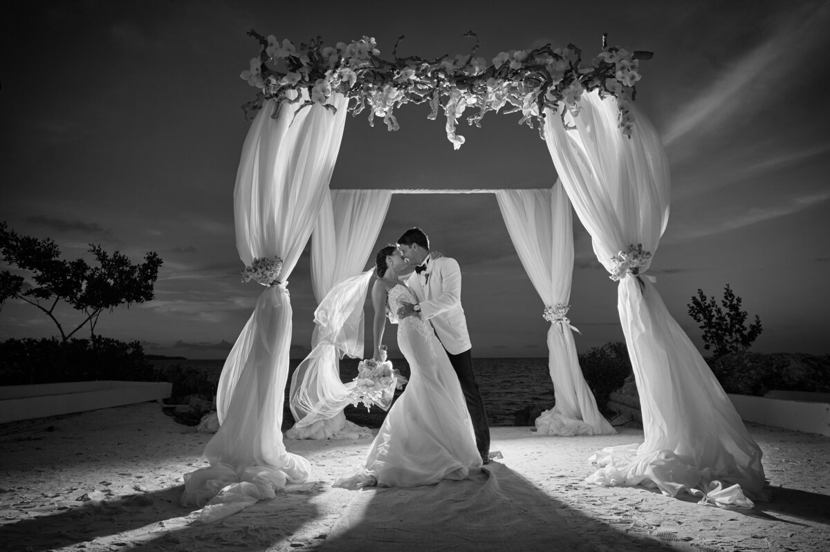 A bride and groom share a kiss under a beautiful, illuminated wedding arch on the beach, captured by Rodrigo Varela Photography.