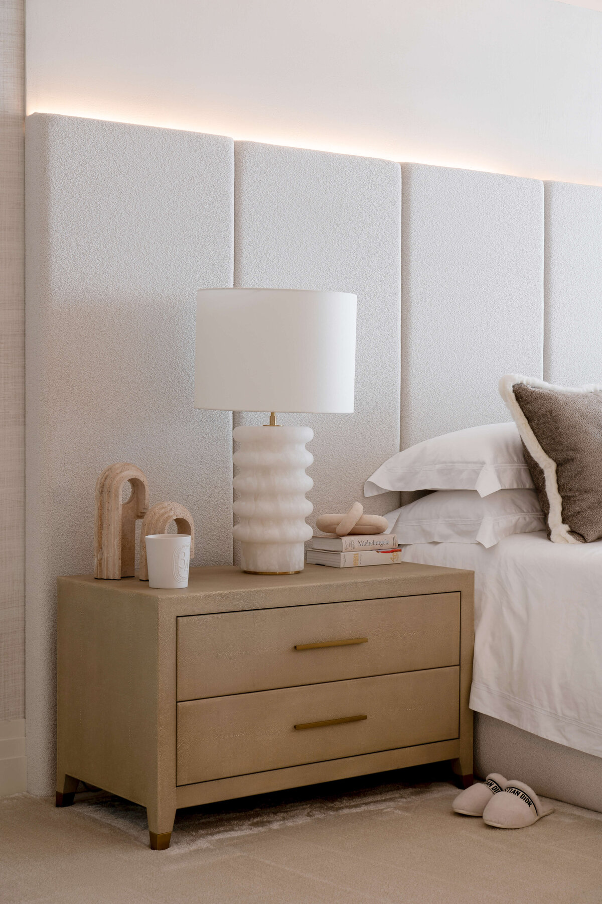 A light tan bedside table next to the primary bed is adorned with gold handles and neutral decor on top.