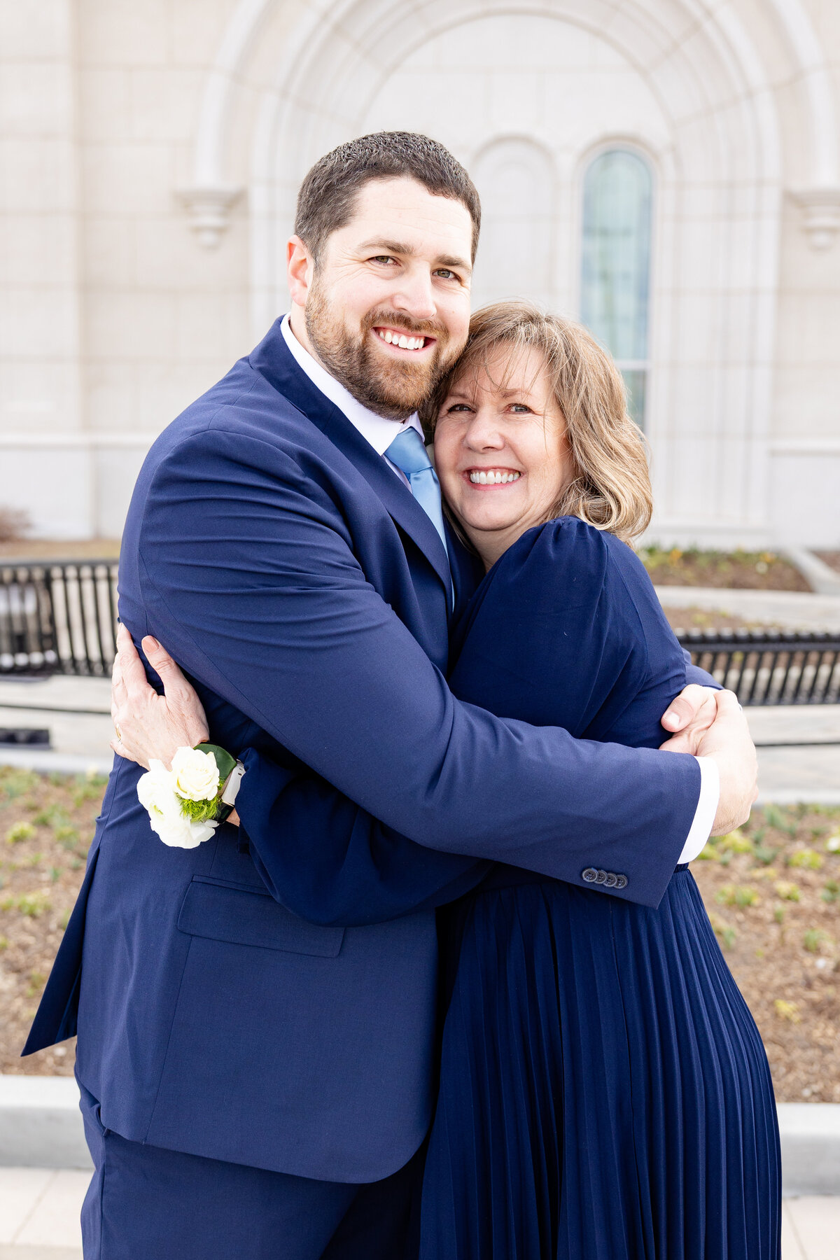 Groom and his mother share a joyful hug, smiling at the camera while standing outside the temple, with flower corsages visible.
