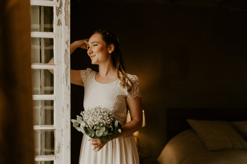 Mariée souriante avec son bouquet accoudée à une fenêtre ouverte, observant l'extérieur immortalisée par Laura, photographe mariage en vendée.