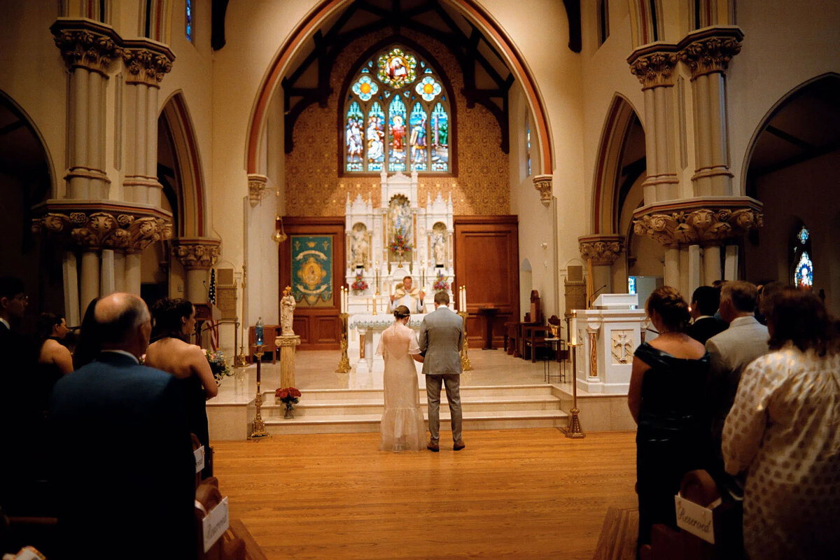A bride and groom stand at the altar in a church during their wedding ceremony, facing a priest. Guests are seated on both sides, as a film photographer NJ captures the moment amid stained glass windows and ornate columns.