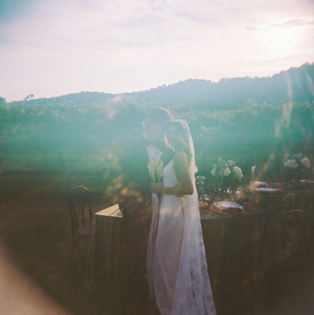 A film image of a bride and groom with a sun flare standing in front of a vineyard.