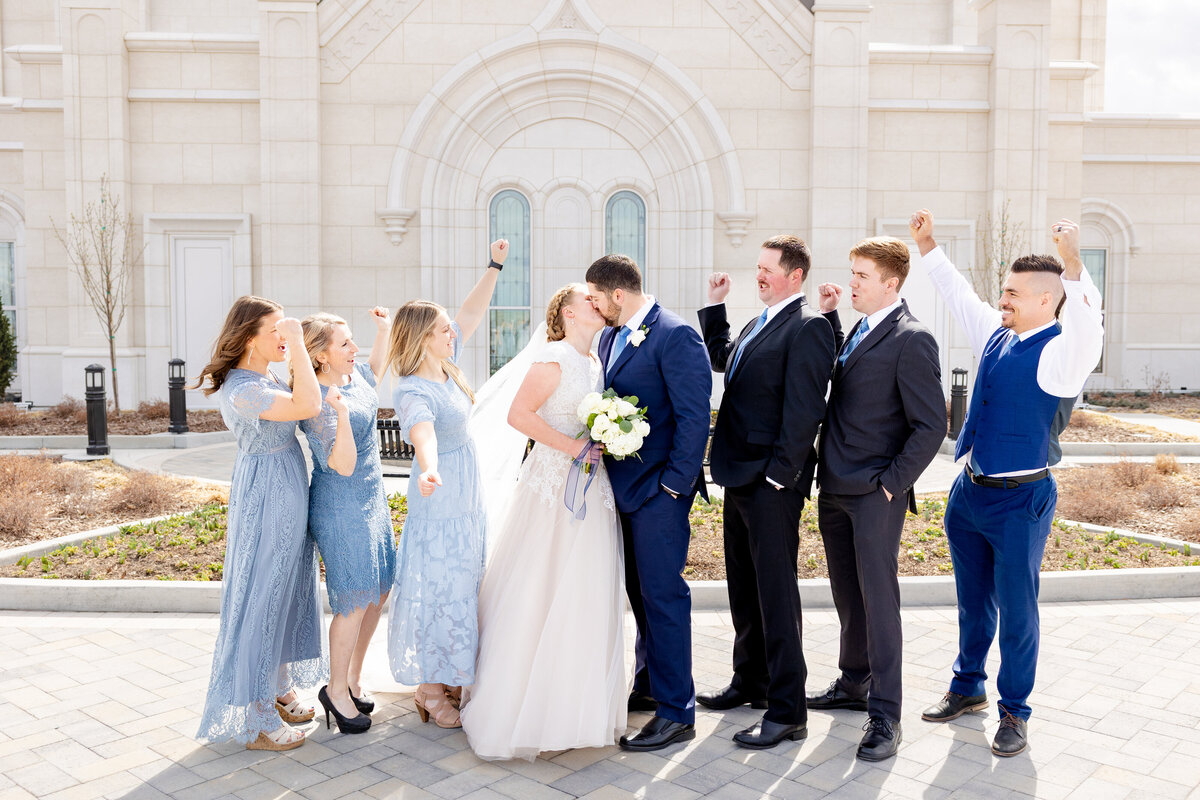The wedding party cheers as the bride and groom kiss joyfully in front of the LDS temple’s arched windows.
