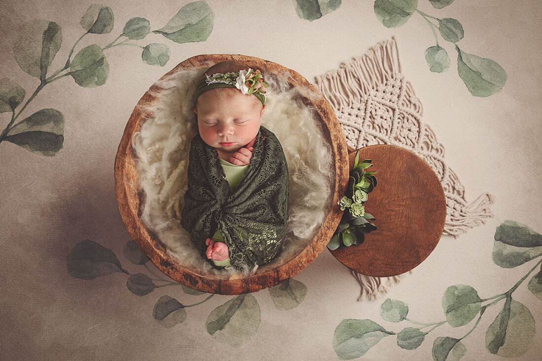 Newborn girl sleeps in a rustic wooden bowl, wrapped in lace with soft greenery and textures.
