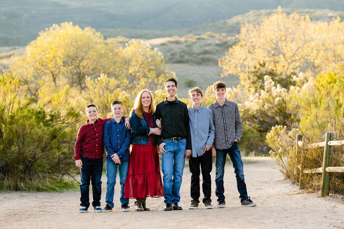 portrait of a family of 2 parents plus 4 boys. They are dressed in plaid and jeans. They are lined up in a dirt trail with plants and trees in the background