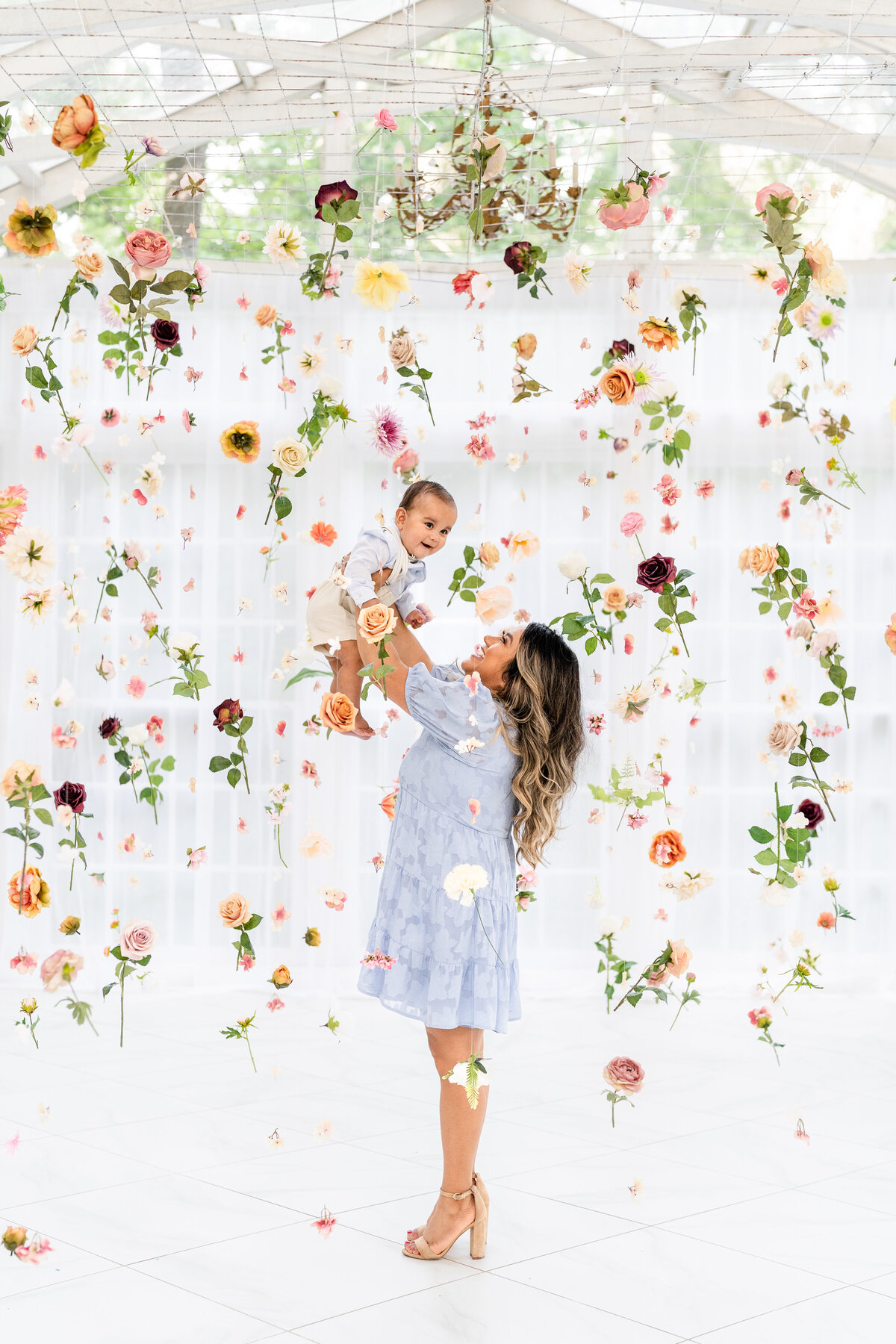 Mom holding baby up in the middle of a hanging flower set in the Willow all white greenhouse  at the Oak Atelier