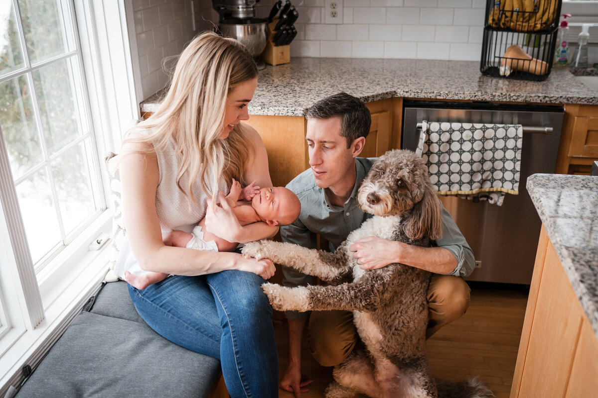A family in their kitchen, the mother holds a newborn baby while the father kneels beside their large dog, who is standing up on its hind legs.