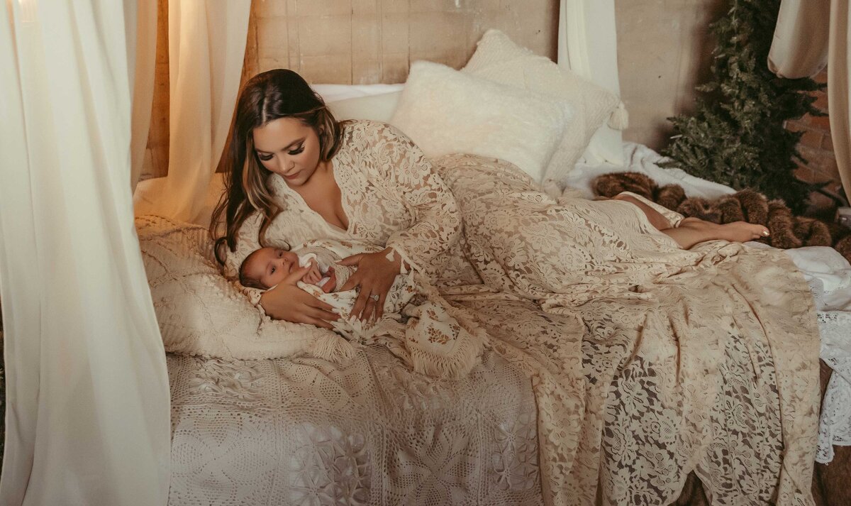 Mother and newborn baby laying on abed. Mother is wearing a long lace dress