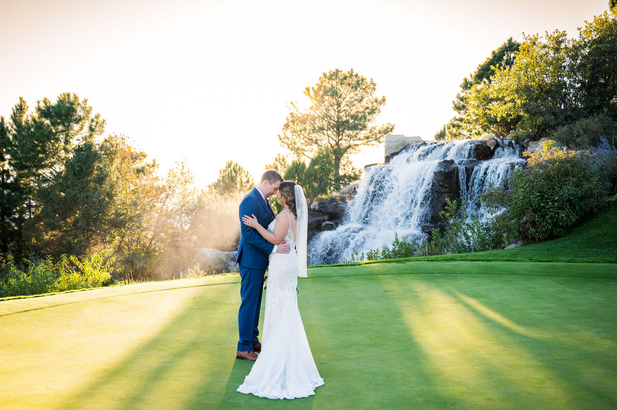 A bride and groom stand forehead to forehead on a golf course with a waterfall in the background.