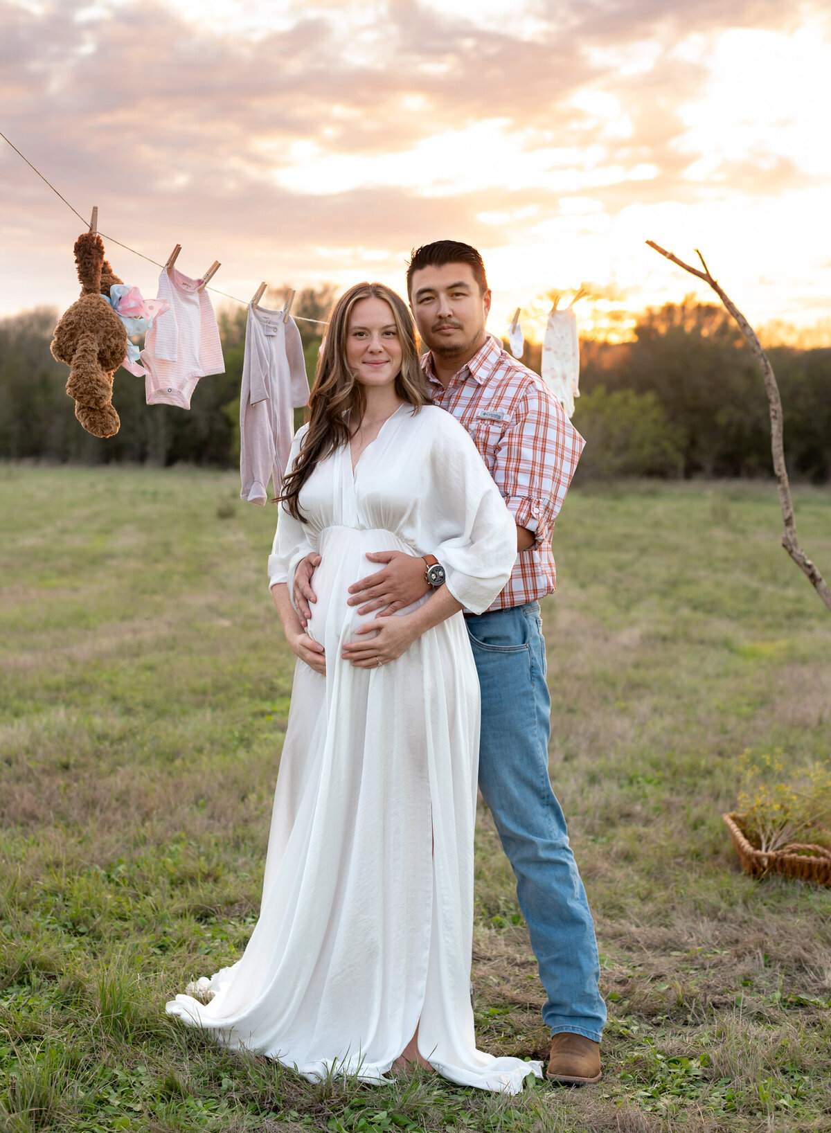 Pregnant couple standing in a field under a laundry line of baby clothes at sunset in Liberty Hill, Texas