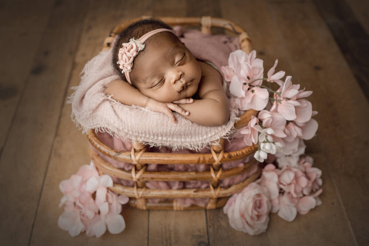 african american baby posed with head on hands in bamboo basket surrounded by pink florals, niagara, ON newborn photography studio