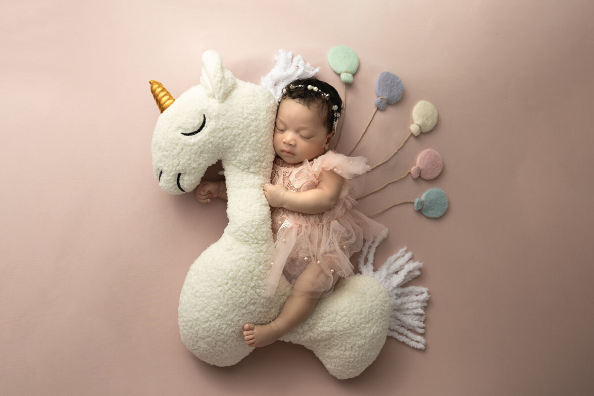A baby rides on the back of a stuffed unicorn holding colorful balloons.