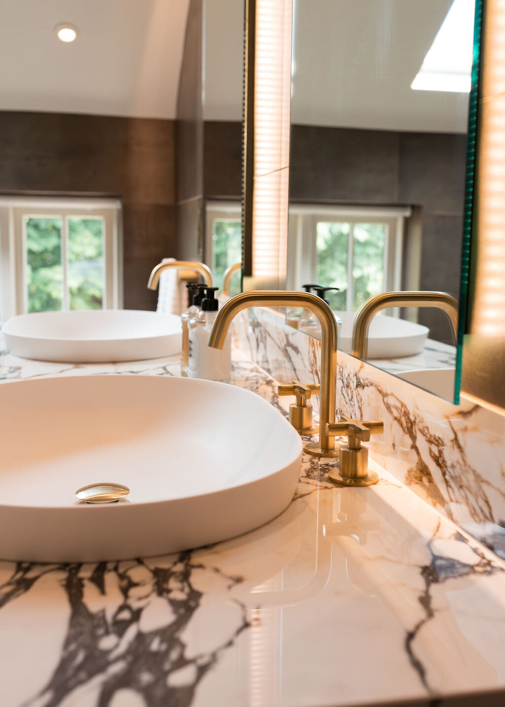 A modern bathroom with a double sink vanity with marble countertops. The sinks feature a white bowl and shiny gold hardware.