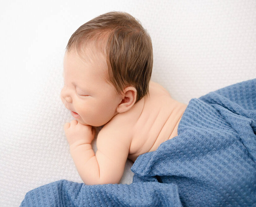 Baby newborn boy sleeping with blue waffle blanket over him while lying on white posing blanket.