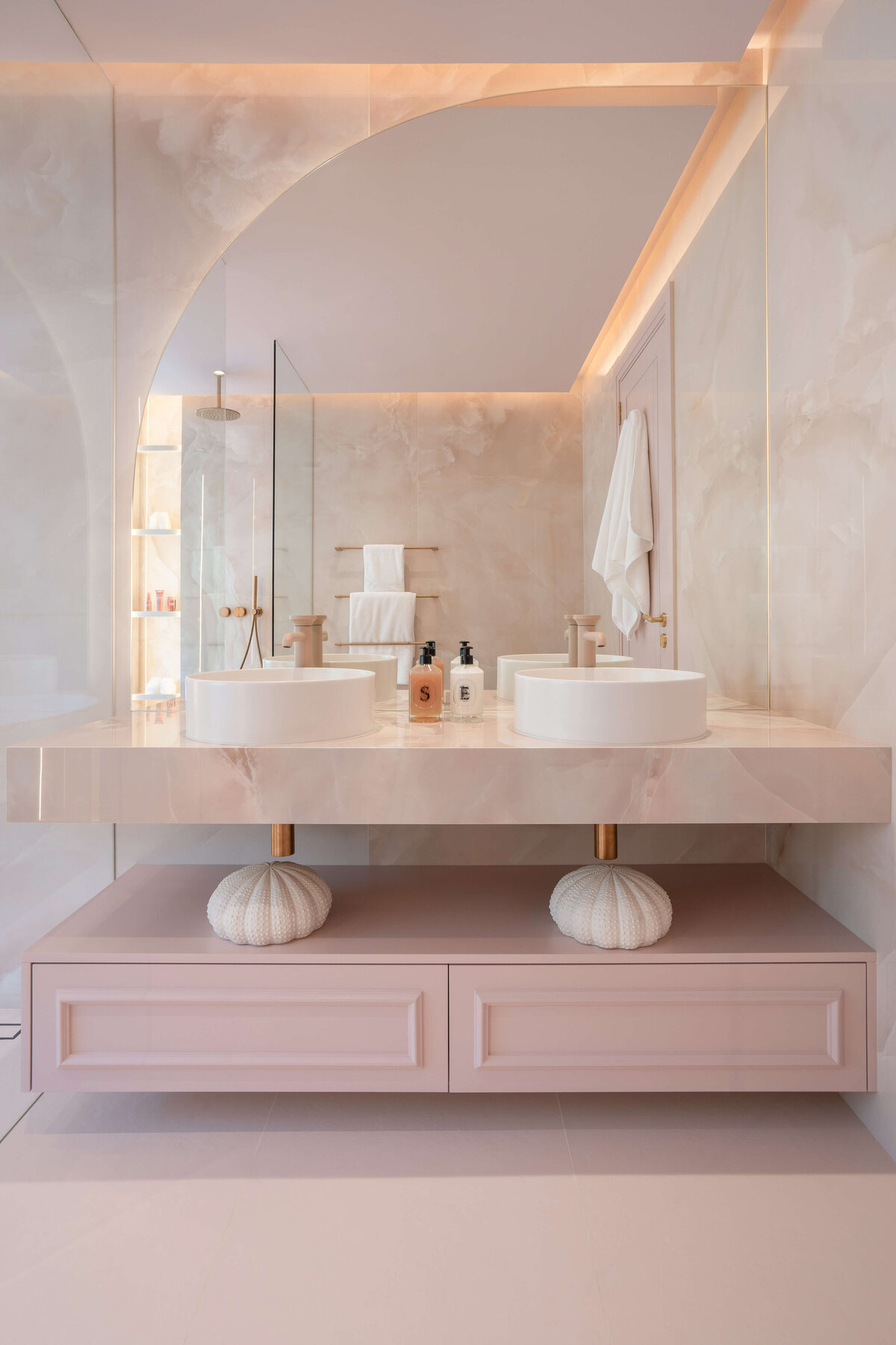 Double sinks in a light pink marble bathroom. The cabinets are pink, the counters marble pink, with matte pink spouts and white bowls. The mirror above the two sinks has one rounded  corner at the top left.