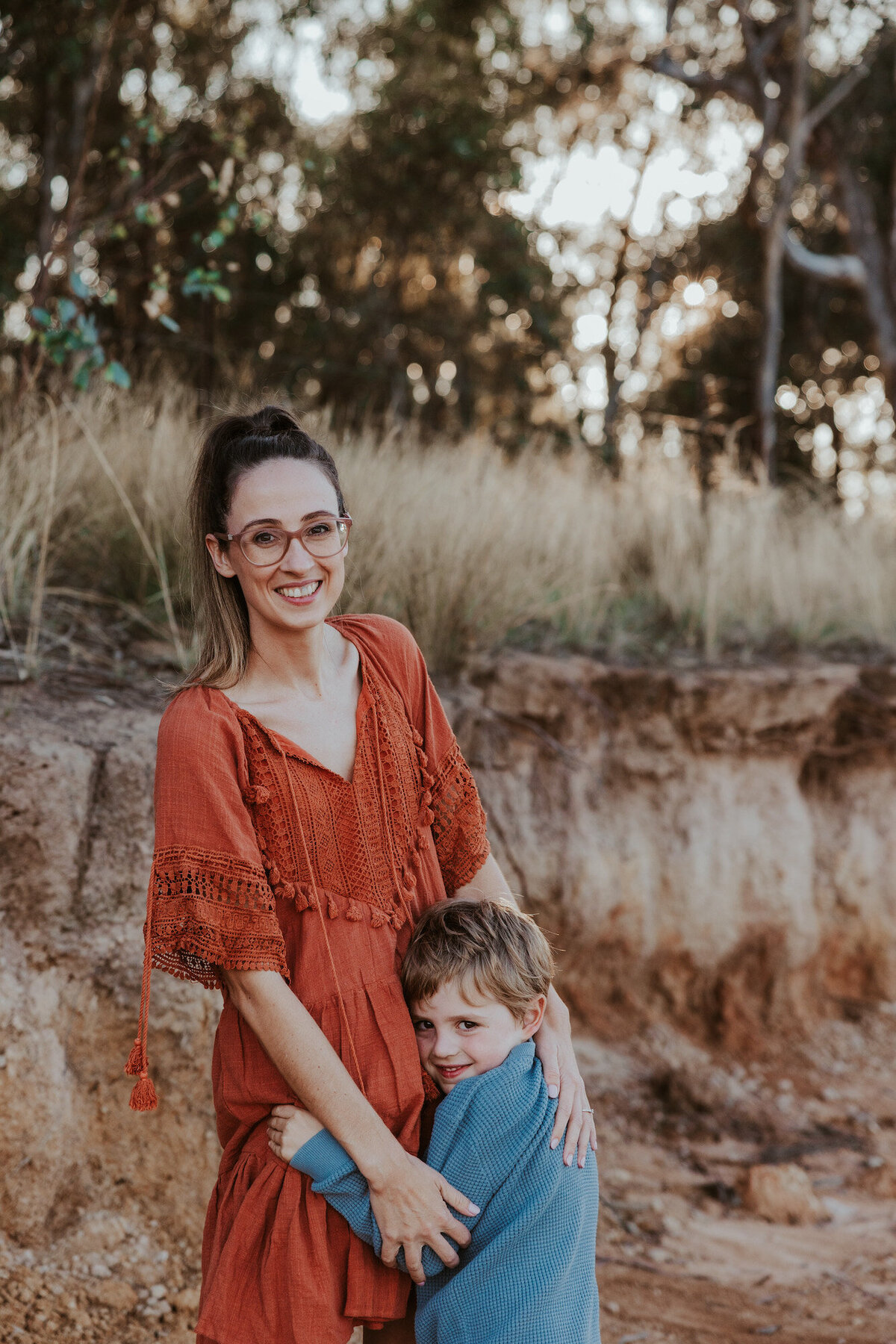 Mum smiling at the camera while her son snuggles into her shoulder, cheekily grinning with his back turned during a family photo shoot