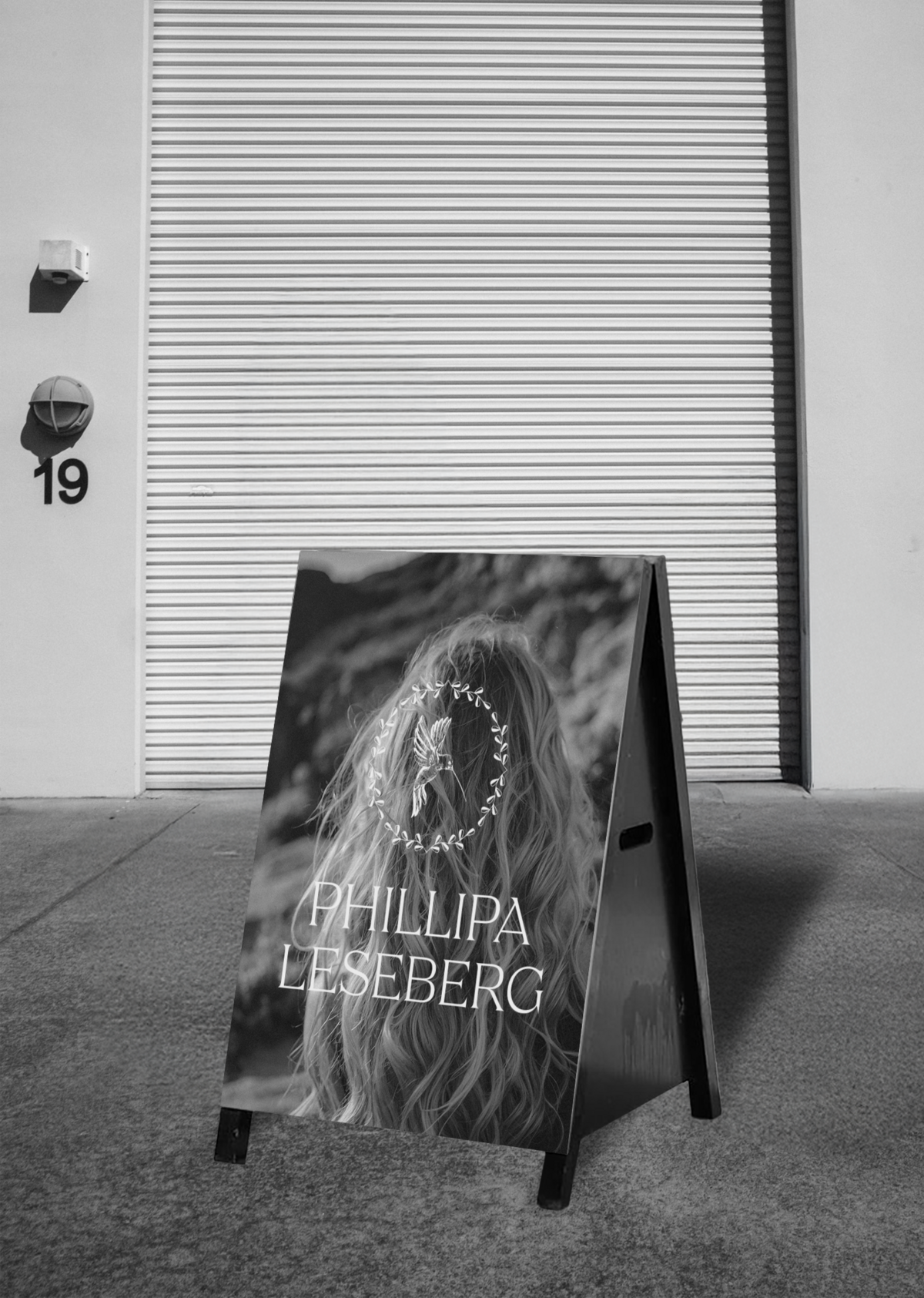 A sandwich board sign with the text "PHILLIPA LESEBERG" and an image of a woman with long hair stands on a sidewalk in front of a closed industrial garage door marked with the number 19, showcasing a brand designer client mockup for Sweet Pea Ink Creative.