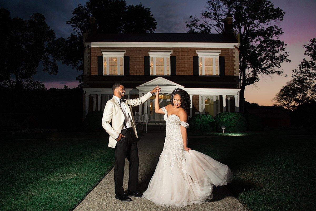 The groom, dressed in a black and white  tuxedo, spins the bride, dressed in a white lace mermaid style dress as they dance at sunset in front of Ravenswood Mansion