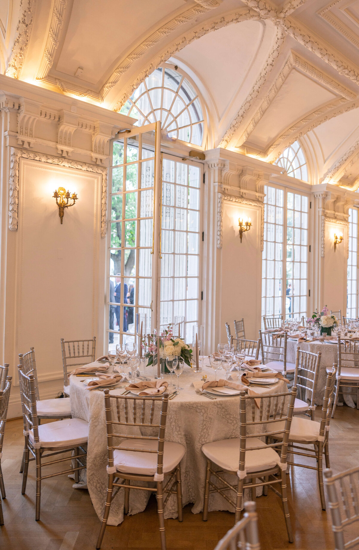 Elegant banquet hall with arched windows and ornate molding. Tables are set with white tablecloths, silver Chiavari chairs, and floral centerpieces. Soft lighting creates a warm, inviting atmosphere.