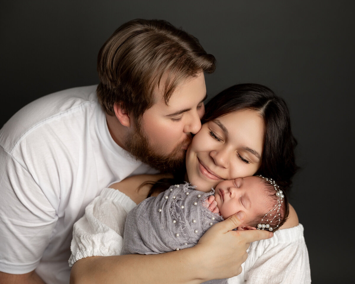 A loving father kissing the cheek of his wife as they both gaze down at their peacefully sleeping newborn daughter, wrapped in a pearl-encrusted gray swaddle and adorned with a matching headband. Captured by Inland Empire newborn photographer Kaitlyn Dawn Photography.