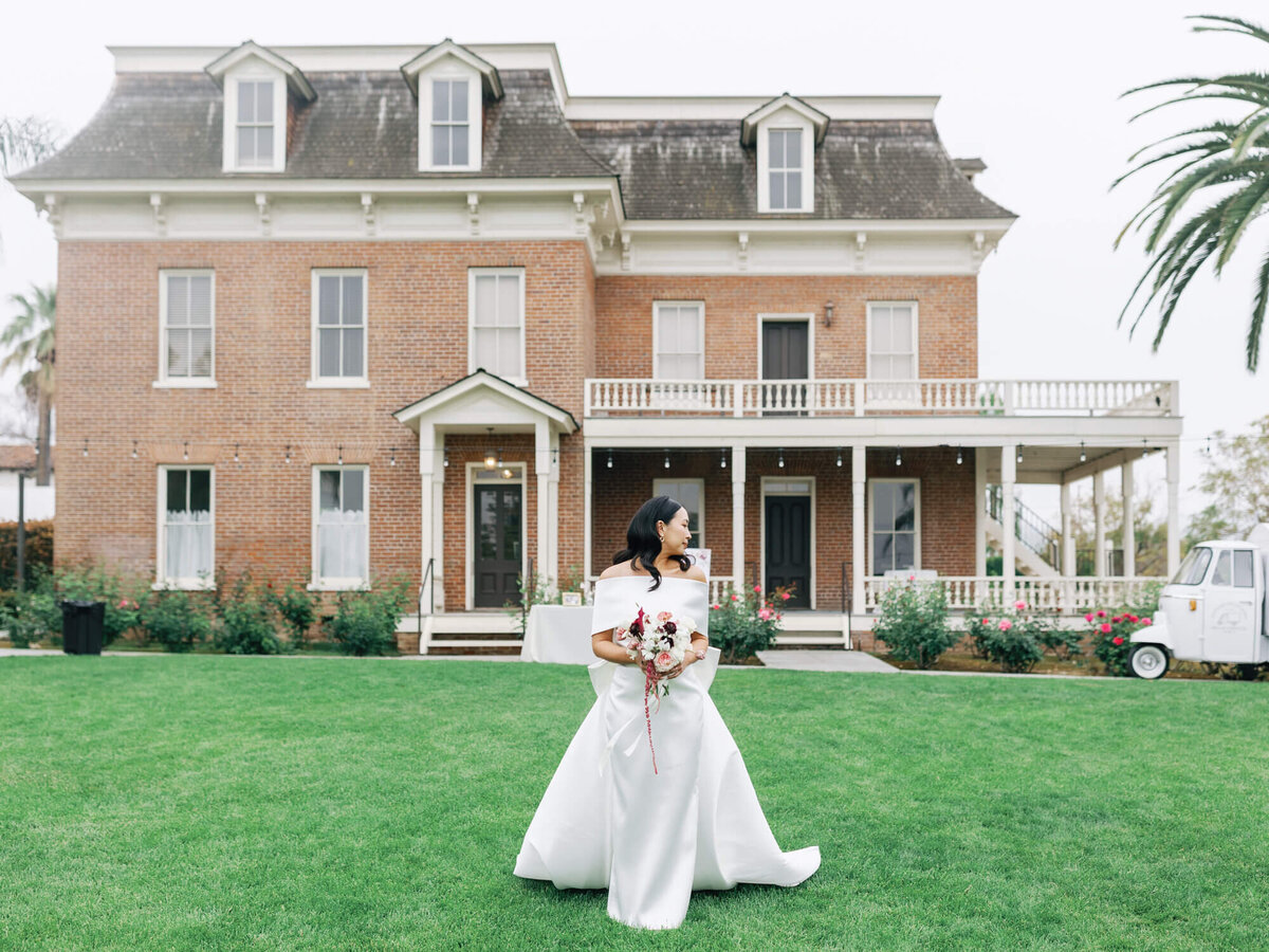A bride in a white gown stands on a lawn, holding a bouquet and smiling, with a large Victorian-style brick house in the background.